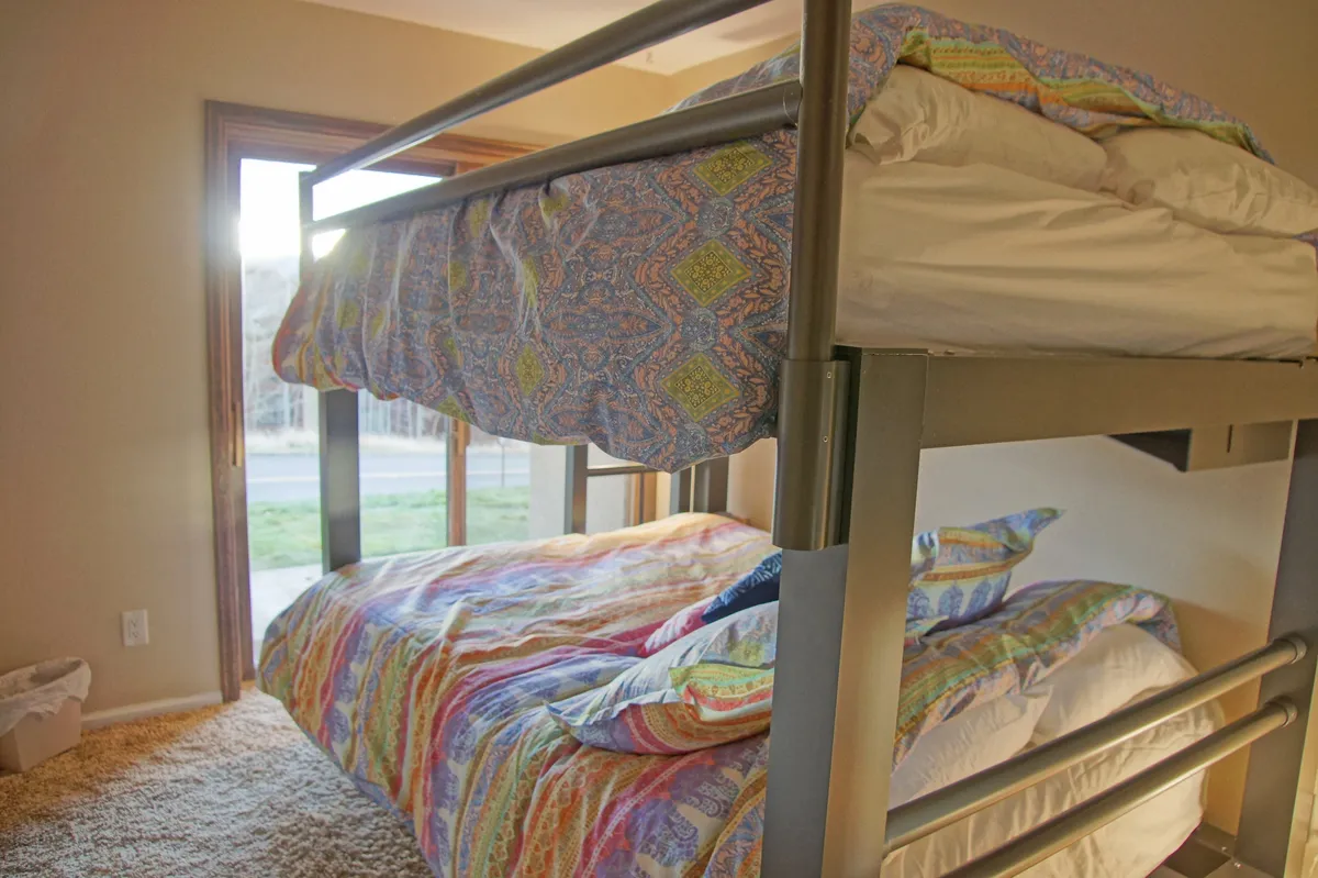 A cheerful bedroom with a bunk bed dressed in colorful bedding, complemented by a window that brings in abundant morning sunlight and offers a glimpse of the surrounding landscape. The space feels playful yet restful, inviting shared stories and relaxation after a day of adventure.