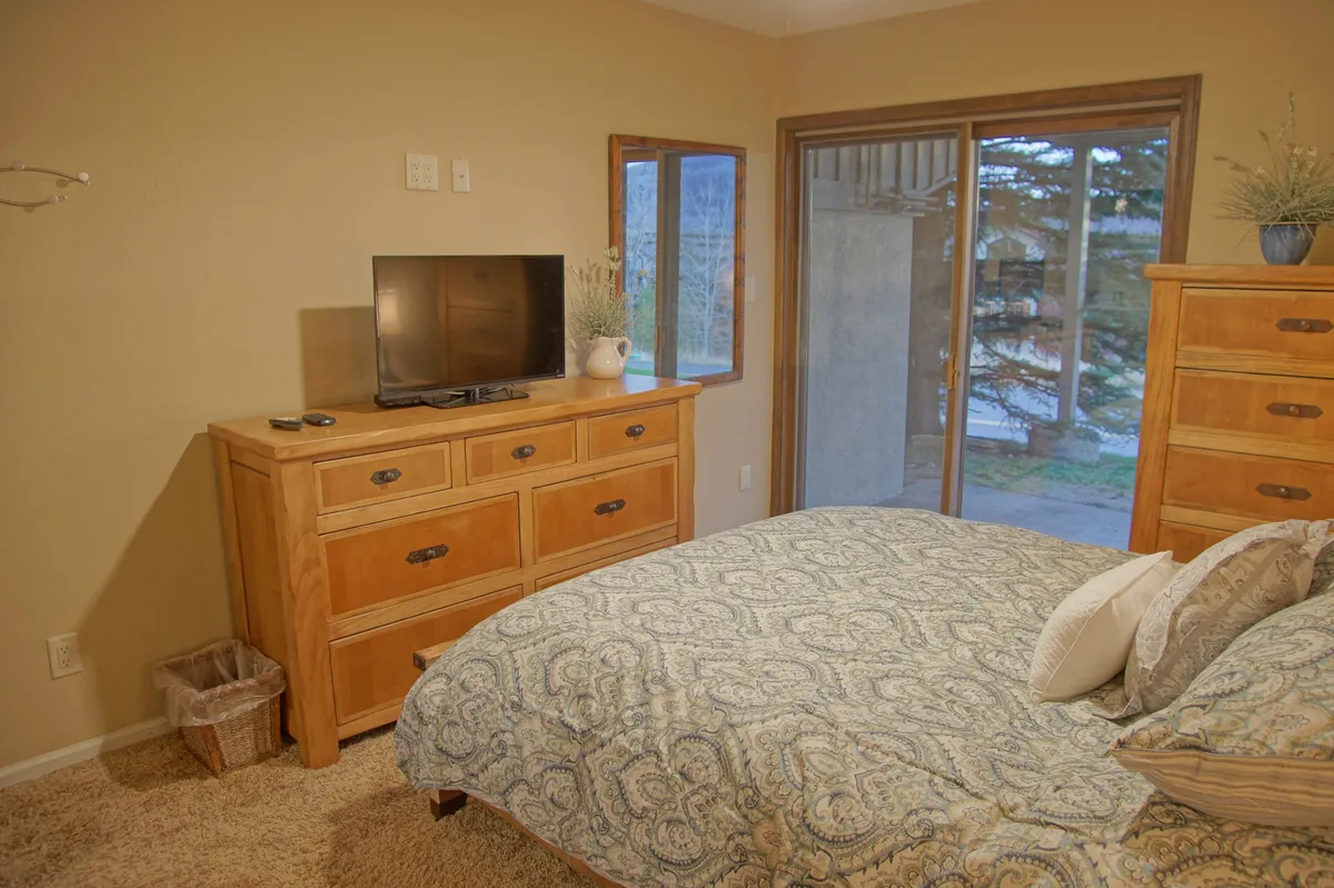 A spacious bedroom filled with natural light from large windows. The setup includes a comfortable bed, dresser, and a television, creating an inviting space for both relaxation and entertainment. The window view and the overall brightness hint at a homey atmosphere ideal for family stays or remote work.