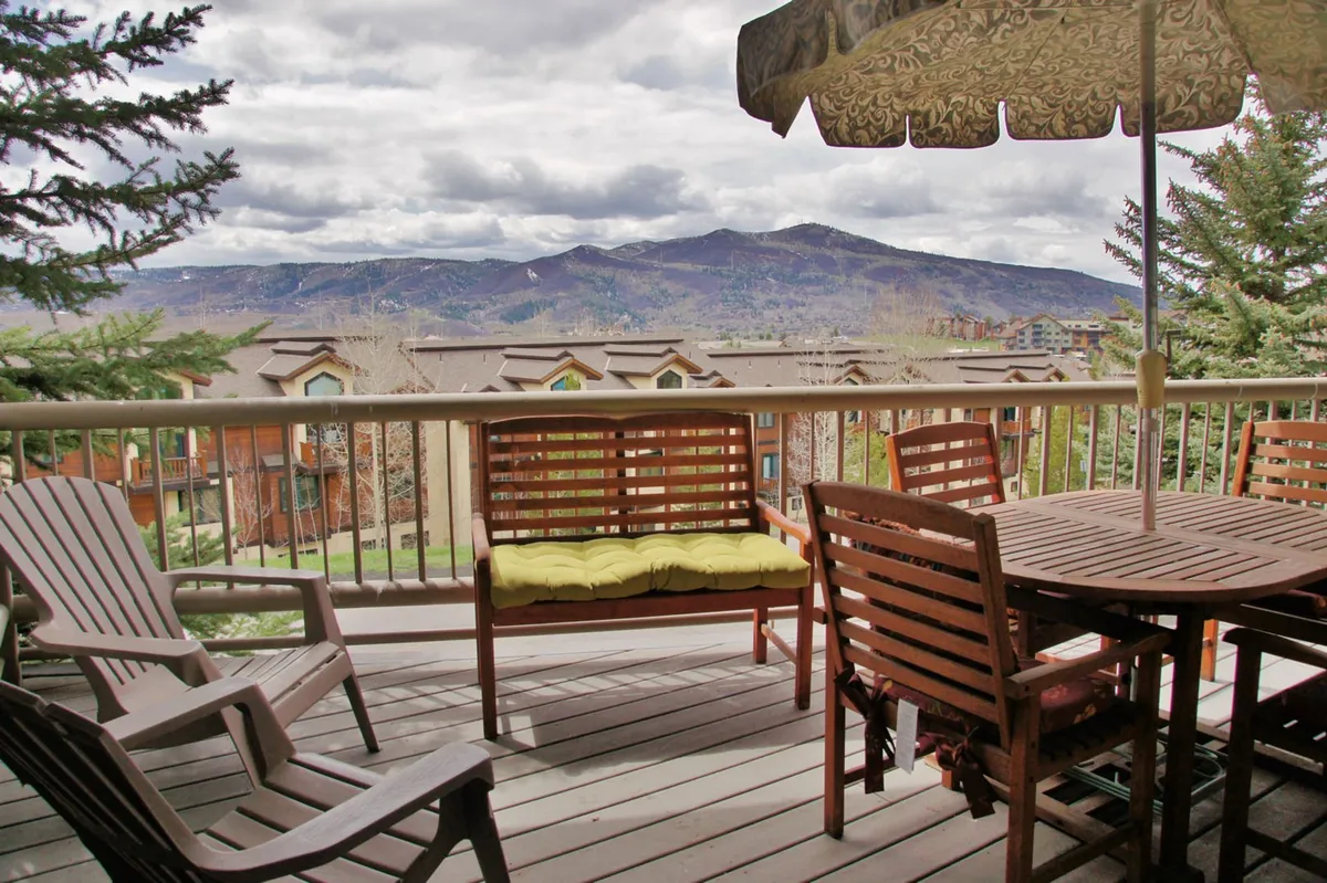An inviting deck is furnished with comfortable outdoor seating and umbrella-shaded tables, all oriented toward a breathtaking mountain view. The ambiance is tranquil, perfect for enjoying a sunset, dining outdoors, or simply relaxing with friends and family.