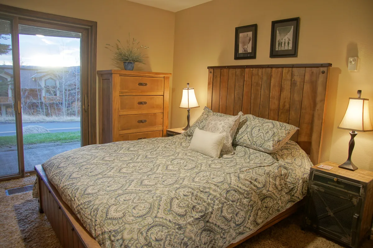A warmly lit bedroom featuring a wooden bed frame with plush decorative pillows, a lamp casting a cozy glow on the bedside table, a chest of drawers, and tasteful framed pictures on the wall. Through the window, sunset light spills into the room, hinting at beautiful valley views and tranquil evenings.