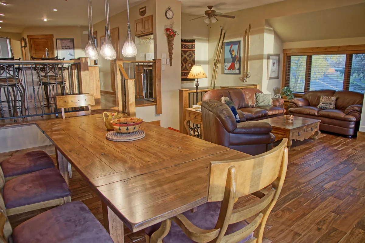 An open-plan living and dining area showcasing a large wooden dining table set for family meals, comfortable leather sofas, pendant lighting, and decorative wall art. The layout encourages communal dining, storytelling, and cozy relaxation by the fire.