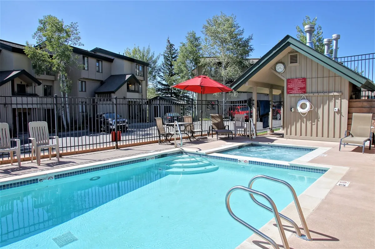 The image shows a sparkling heated swimming pool surrounded by inviting lounge chairs under a vibrant red umbrella, set against a backdrop of Colorado mountain scenery at sunset. The scene evokes a sense of relaxation and leisure, perfect for après-ski swims, poolside barbecues, and gathering with friends or family.