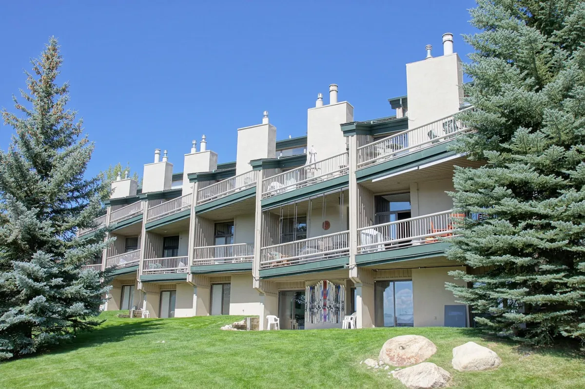 This exterior image showcases the property's balconies overlooking a green lawn, framed by tall pine trees and striking rock formations. The scene conveys a calm, nature-focused atmosphere, with people relaxing on balconies and socializing outdoors, emphasizing the property's proximity to outdoor activities.
