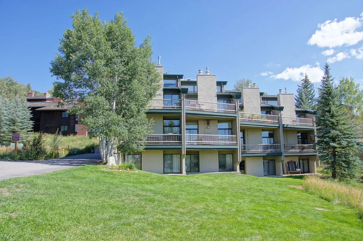 A wide shot of the building's facade looking out onto a green lawn and surrounded by trees. The balconies suggest communal outdoor spaces, perfect for morning coffee or gathering after a day on the slopes. The tranquil mood underscores relaxation and connection to nature.